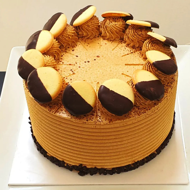 A round coffee cake with Tiramisu, covered in chocolate chips and cookie crumbs, on a white cake stand.